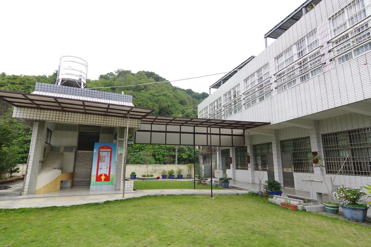 明湖國小的後方庭院：草地、雨遮走廊與洗手台，教室沿著山坡層層排開，後方是覆滿綠樹的山壁 A rear courtyard at Minghu — lawn, a covered walkway and washing troughs, classrooms stepped up along the slope, with the tree-covered hillside directly behind
