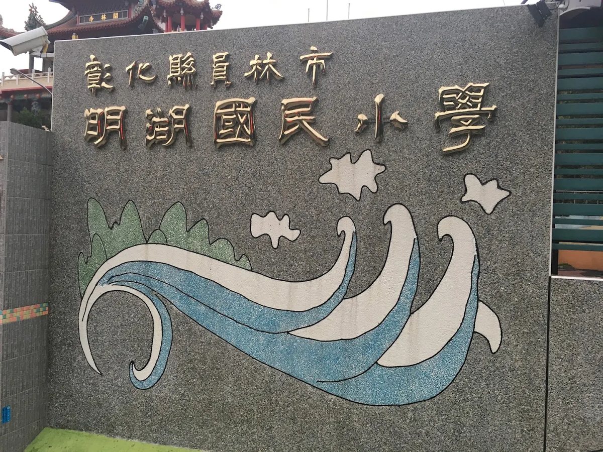 明湖國小的花崗岩校名牆：金色立體字「彰化縣員林市明湖國民小學」，下方是綠色山巒與藍白水波的洗石子浮雕，牆頂後方露出開林寺的紅瓦 The granite school-name wall at Minghu — 彰化縣員林市明湖國民小學 in raised gold characters above a washed-stone relief of green hills and blue-and-white water, with the red tiles of Kailin Temple showing behind the top of the wall