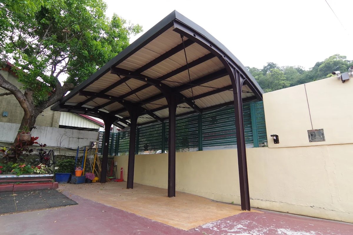 明湖國小新建的戶外活動棚：褐色鋼架、弧形屋頂與採光板，下方是平整的活動空間，一側是綠色格柵圍牆，旁邊有大樹與紅色球場 A newly built outdoor activity shelter at Minghu — a brown steel frame with a curved roof and translucent panels over a level surface, a green louvred wall along one side, a large tree beside it and the red ball court alongside
