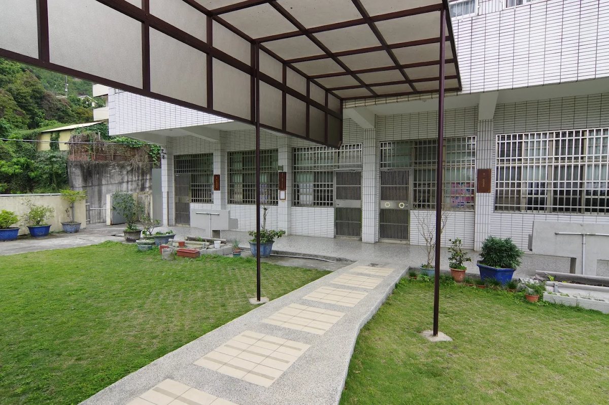 明湖國小的雨遮走廊與教室外的草地：褐色鋼架玻璃頂棚、洗石子步道、走廊上一列盆栽，教室門旁掛著木質班牌 The covered walkway and the lawn outside the classrooms at Minghu — a brown steel frame with a glazed roof, a washed-stone path, a line of potted plants along the corridor and wooden class signs beside the classroom doors