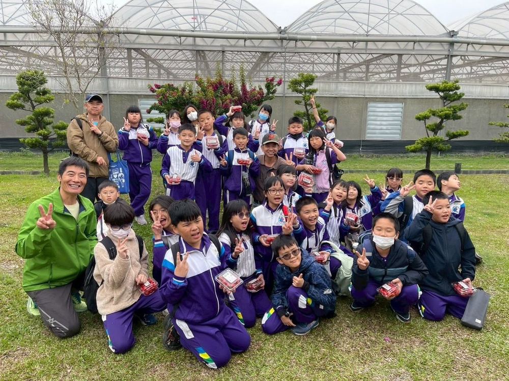 南鎮國小學生在溫室外的草地上大合照，背後是一整排溫室 Nanjen students in a big group photo on the lawn outside a row of greenhouses