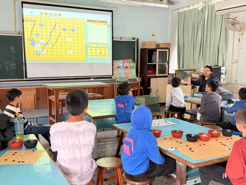 南鎮國小學生在教室裡下圍棋，前方投影幕上是棋盤 Nanjen students playing Go (weiqi) in a classroom, with a game board projected on the screen
