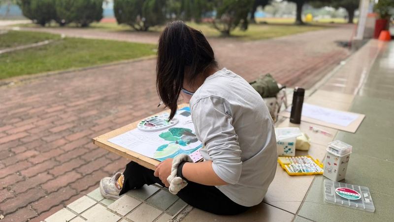 南鎮國小學生坐在校園走廊邊的地上寫生 A Nanzhen student sitting on the ground by a campus walkway, painting outdoors