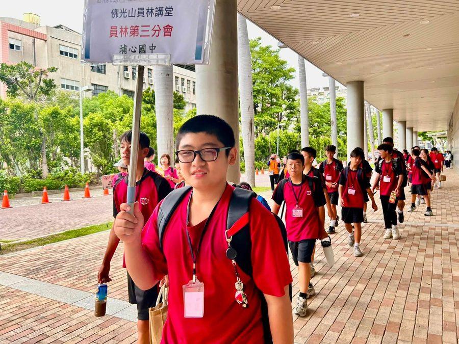 學生手持「佛光山員林講堂・員林第三分會・埔心國中」的牌子，隊伍走在場館外的迴廊 A student carrying a Fo Guang Shan Yuanlin / Pusin JH sign, the group walking along an outdoor arena walkway