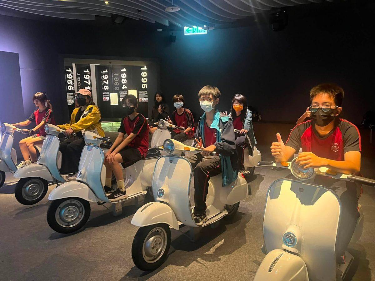 同學騎在中國信託文薈館的偉士牌機車展品上，牆上是 1966、1971、1978 等年代標示 Students on the Vespa scooter exhibits at the CTBC Wen-Hui Hall, a wall of dates behind them