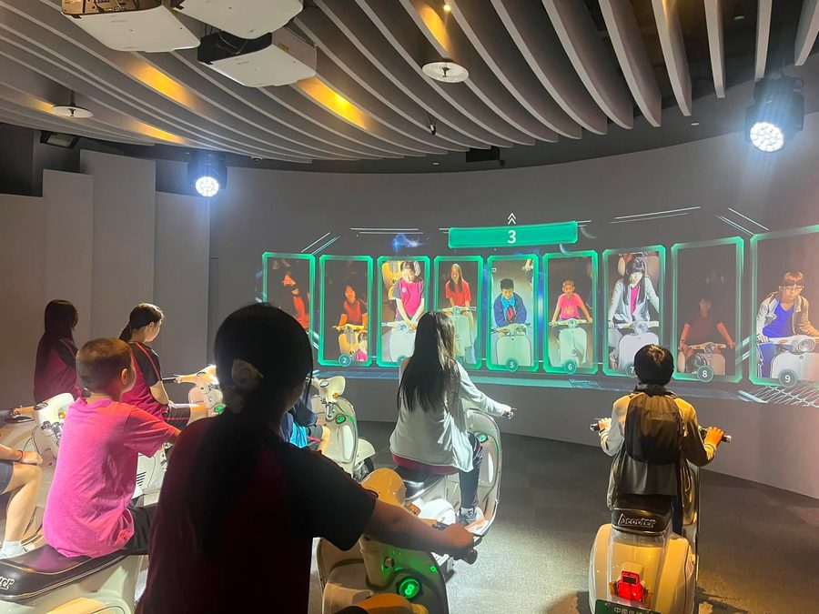 同學坐在機車體驗機上，面對大螢幕上的競速畫面 Students on scooter simulators facing a racing screen