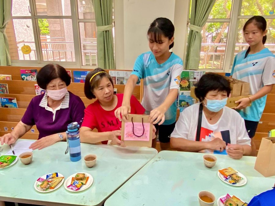 學生雙手把裝著小禮物的紙袋遞給坐在桌邊的阿嬤們 A student handing a gift bag to grandmothers seated at a table