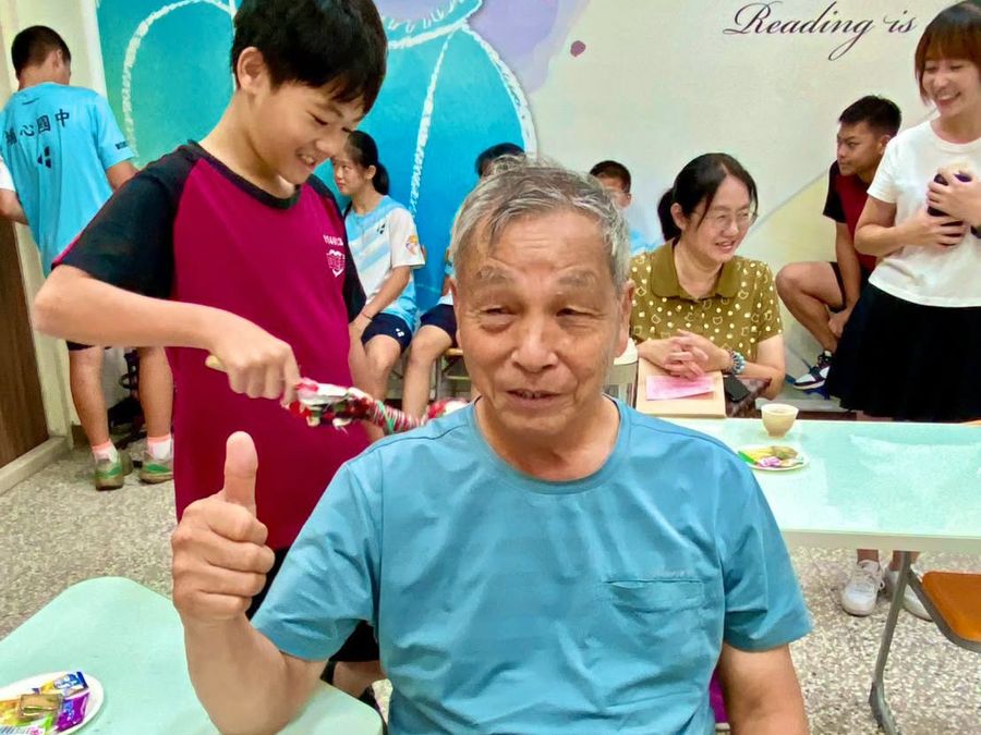 阿公坐著比出大拇指、面帶笑容，身後有學生為他打理頭髮 A grandfather sits smiling with a thumbs-up, a student tidying his hair behind him