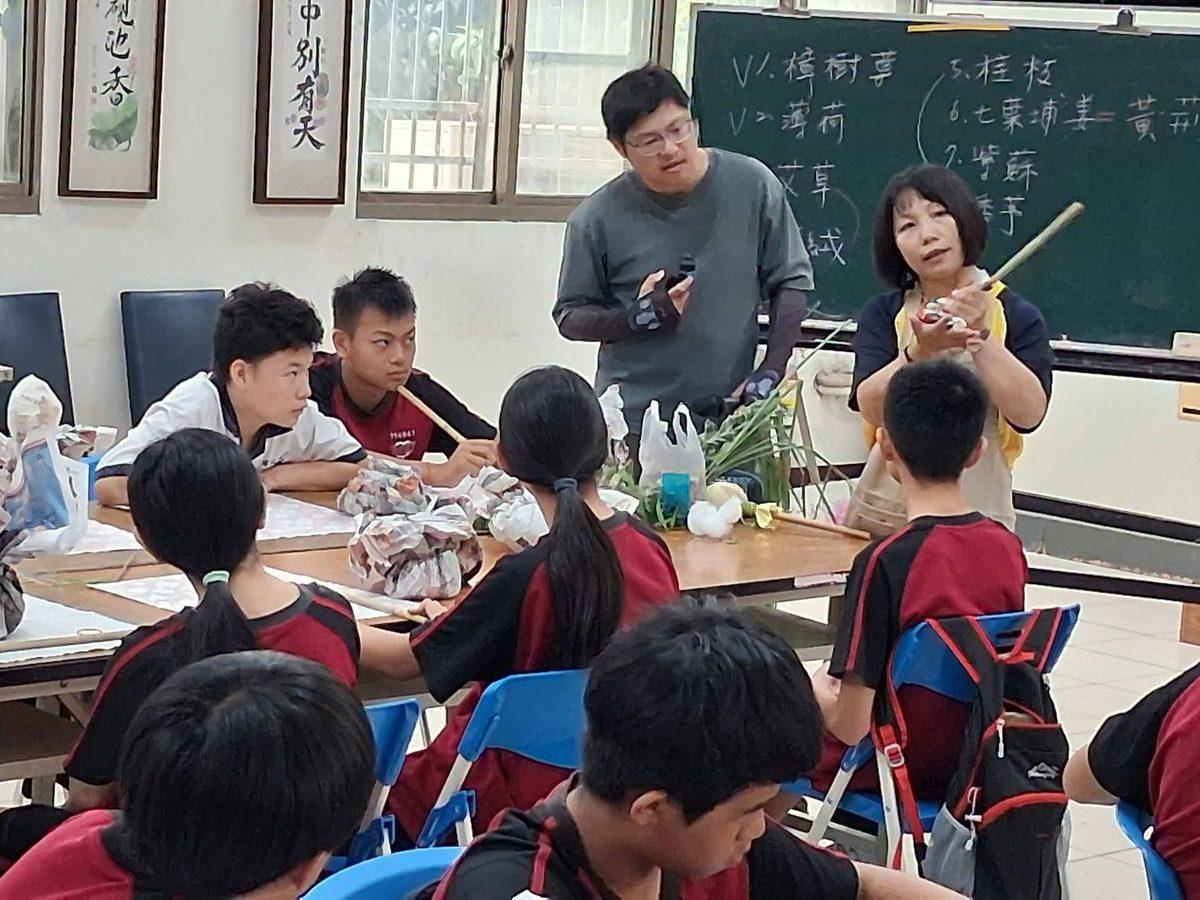 教室裡，老師與志工向坐著的學生講解，黑板上列著樟樹亭、薄荷、艾草、桂枝、七葉埔姜、紫蘇、香茅等香草名稱 In a classroom a teacher and volunteer explain to seated students, a blackboard listing herb names