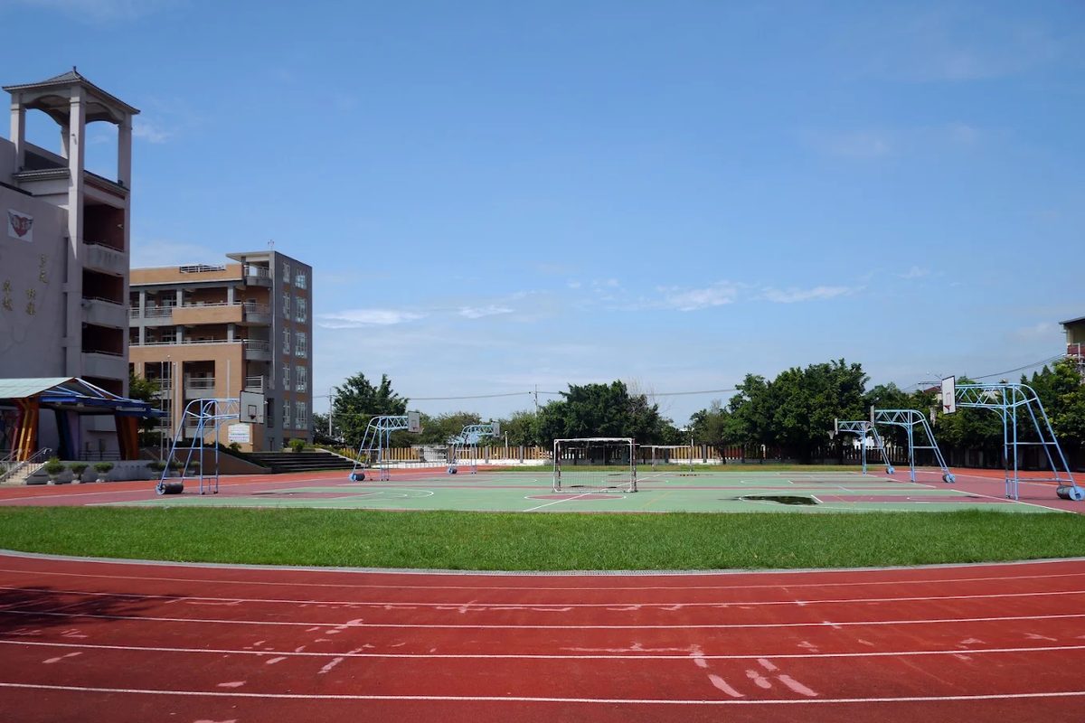 埔心國中的紅色 PU 跑道與綠地球場，遠處是鐘樓校舍與藍天 The red running track and green ball courts at Pusin, the bell-tower building and a blue sky behind