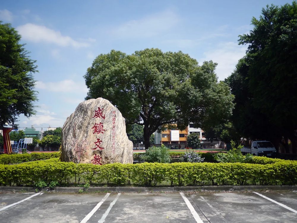埔心國中停車場旁一塊刻著「威鎮文教」的巨石，後方是一棵大樹與藍天 A large boulder in the Pusin grounds carved with the characters for 'a stronghold of culture and learning', a big tree and blue sky behind