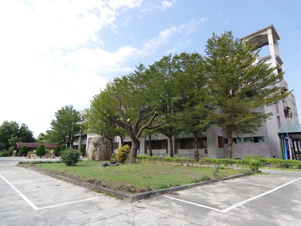 埔心國中校園裡的老樹與鐘樓校舍，樹下是整齊的停車格與藍天 Mature trees and the bell-tower building at Pusin, tidy parking bays beneath them and a blue sky