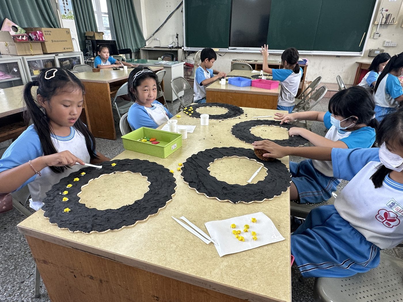 橋頭國小紙黏土社在教室創作：學生們圍著兩張併起來的木桌坐著，桌面上放著四件大型圓環狀的作品，外圈是深色的紙黏土、內圈是米色底板，孩子們正用手把一顆顆黃色的小黏土黏到外圈上；桌上另有裝顏料的白色小杯與綠色置物籃，背景是黑板、窗戶與堆放教具的桌子 The paper clay club at Qiaotou at work in a classroom — children seated around two wooden tables pushed together, four large ring-shaped pieces laid out on the surface with dark clay rims around pale centers, small yellow clay shapes being pressed onto the rims by hand, white pots of paint and a green tray between them, and a blackboard, windows and tables of materials behind