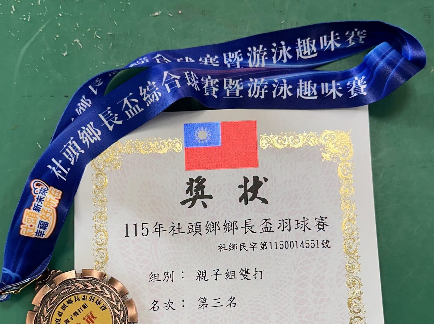 一張獎狀與一面銅牌放在綠色桌面上：獎狀上方印著國旗與金色花邊，中央毛筆字寫著「獎狀」，下方印著「115年社頭鄉鄉長盃羽球賽」，再下面兩行寫著「組別：親子組雙打」「名次：第三名」；左側是一條藍色綬帶，上面重複印著「社頭鄉鄉長盃綜合球賽暨游泳趣味賽」，綬帶末端垂著一面刻字的銅色獎牌 An award certificate and a bronze medal laid on a green table — the certificate headed by the national flag inside a gold border with 獎狀 in brush characters, the line 115年社頭鄉鄉長盃羽球賽 beneath it, and two lines reading 組別：親子組雙打 and 名次：第三名; a blue ribbon printed repeatedly with the name of the township tournament runs down the left side with an engraved bronze medal hanging from it