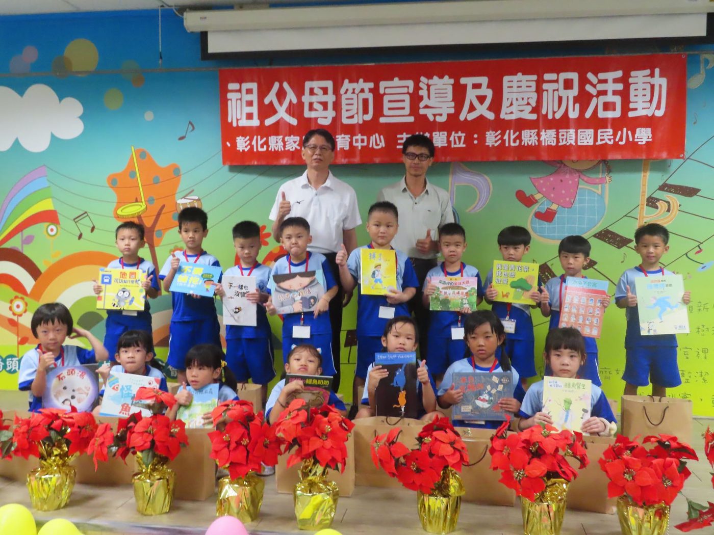 橋頭國小祖父母節活動大合照：紅色布條上寫著「祖父母節宣導及慶祝活動」與「主辦單位：彰化縣橋頭國民小學」，布條下站著兩位老師與十幾位穿藍白運動服的新生，每個孩子手上都舉著一本剛拿到的童書，舞台前擺著一整排包著金色紙的聖誕紅盆花與紙袋 A group photo at Qiaotou beneath a red banner reading 祖父母節宣導及慶祝活動 and naming the school as organizer — two teachers and more than a dozen new pupils in blue-and-white PE kit, each holding up a newly given picture book, with a row of gold-wrapped poinsettias and paper gift bags along the front of the stage
