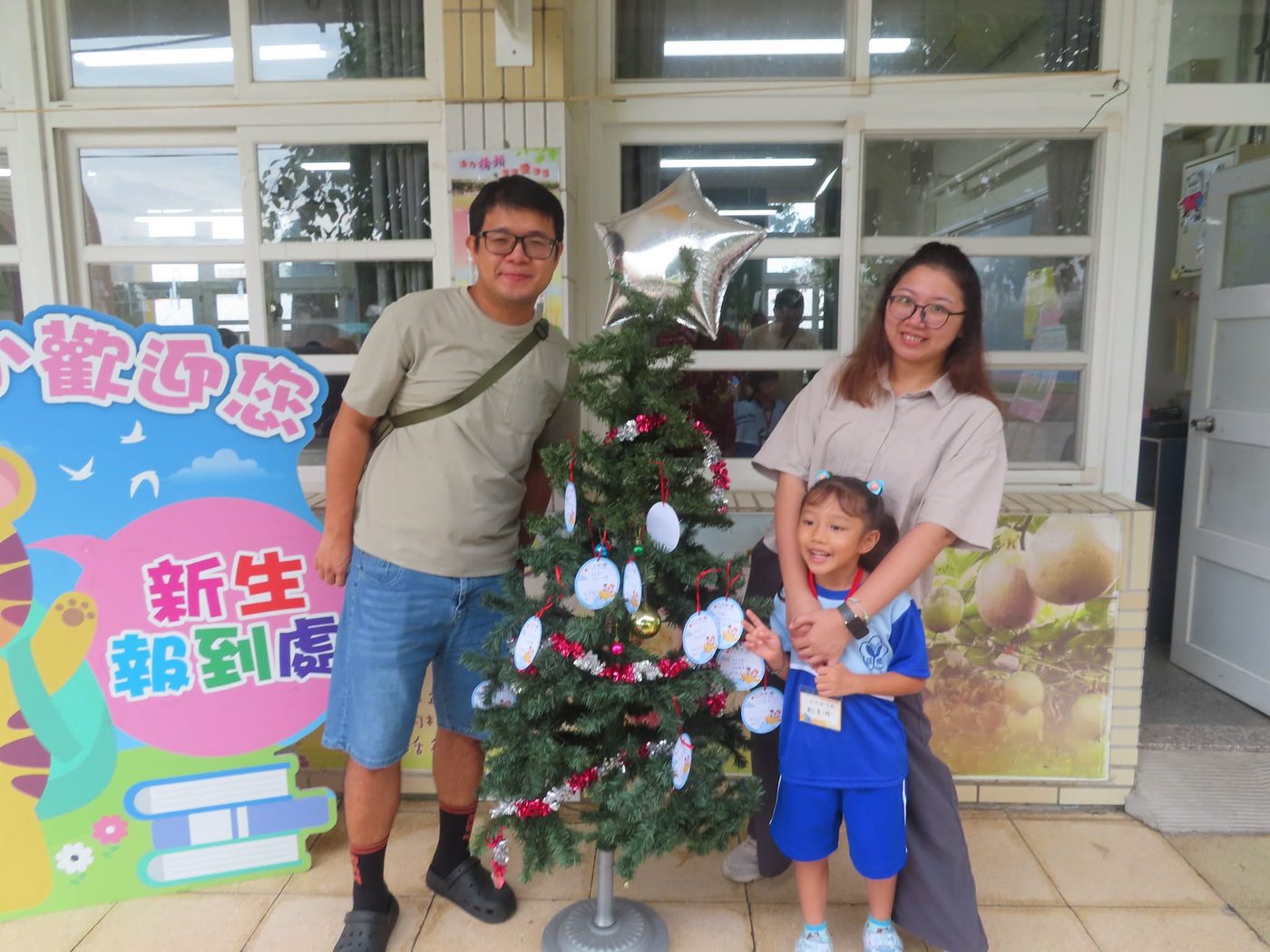 橋頭國小新生報到處：走廊上立著一塊粉藍色的立牌，寫著「歡迎您」與「新生報到處」，旁邊擺著一棵掛滿白色祈願卡與銀色星形氣球的小樹，一位爸爸、一位媽媽和一個穿藍色運動服、比出勝利手勢的小女孩站在樹的兩邊 The registration point at Qiaotou — a pale blue standing board in the corridor reading 歡迎您 and 新生報到處, a small tree beside it hung with white wish cards and a silver star balloon, and a father, a mother and a small girl in blue PE kit making a V-sign standing on either side of it
