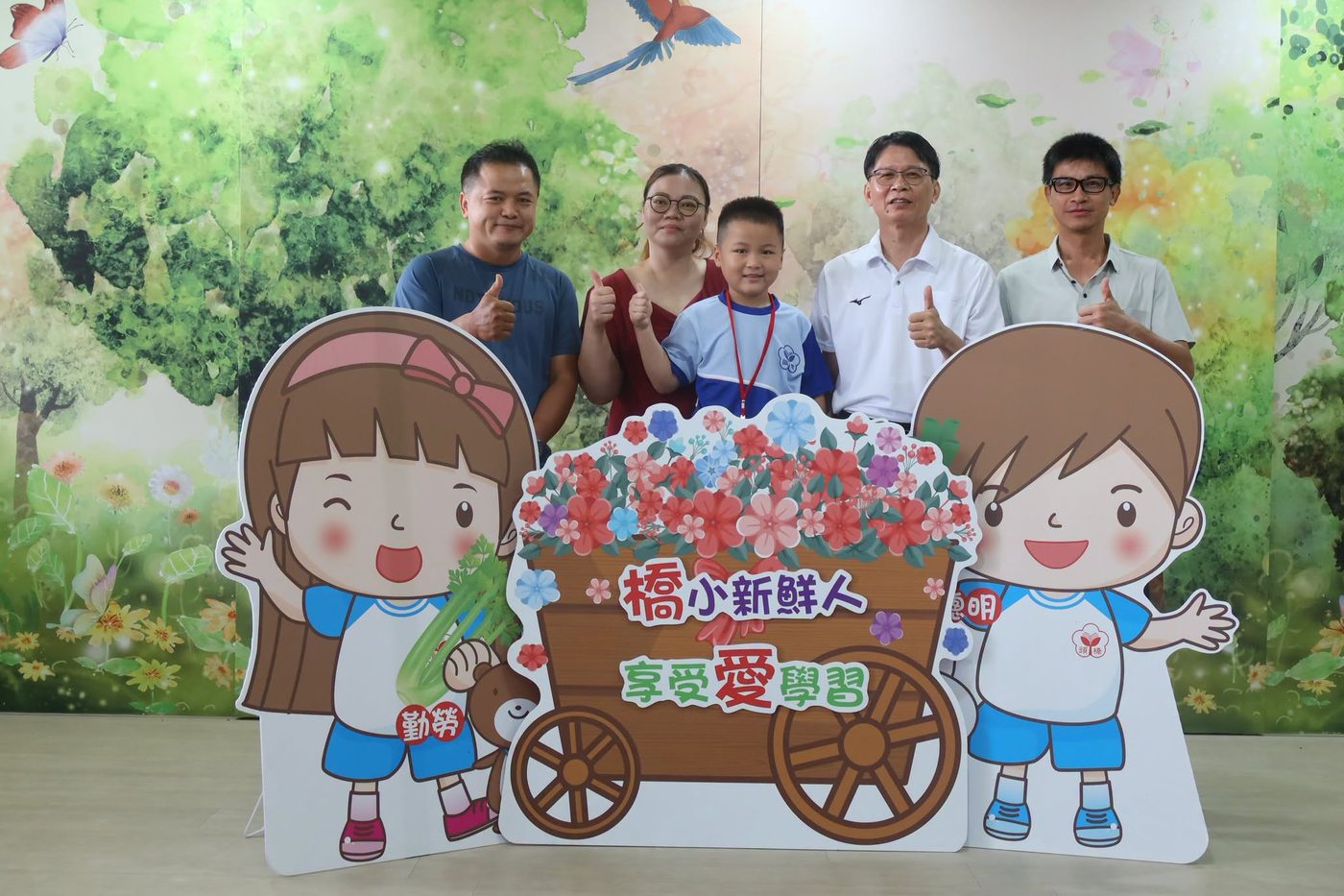 橋頭國小新生打卡合照：一組立體紙板拍照看板立在室內，中央畫著一台裝滿花的木頭推車，車身寫著「橋小新鮮人」與「享受愛學習」，兩側各站著一個卡通學童立牌；立牌後面站著一位穿藍白運動服的男孩、他的父母，以及穿白色 POLO 衫的校長和一位老師，大家比出讚的手勢，背景是綠色森林與飛鳥的彩繪牆 A photo-booth shot at Qiaotou — a cut-out board of a wooden cart full of flowers lettered 橋小新鮮人 and 享受愛學習, flanked by two cartoon schoolchildren; behind it stand a boy in blue-and-white PE kit with his parents, the principal in a white polo shirt and a teacher, all giving a thumbs-up, against a painted wall of green woodland and flying birds