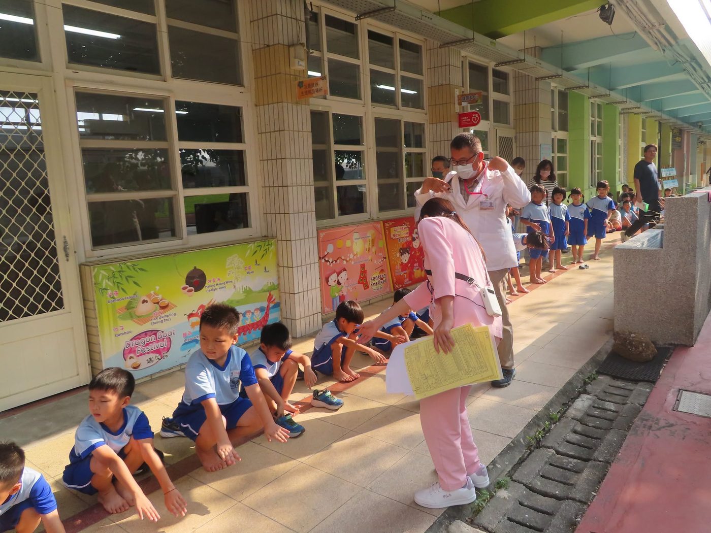 橋頭國小孩子在走廊上等候健康檢查：幾個孩子脫了鞋坐在地上排隊等待，一位穿粉紅色護理服的護理師手裡拿著一疊黃色的健康檢查紀錄卡走過隊伍，後方一位穿白袍的醫師正俯身替孩子檢查，走廊牆上貼著端午節與新年的中英雙語節慶海報 Children waiting their turn at Qiaotou — several of them sitting on the walkway with their shoes off, a nurse in pink scrubs carrying a stack of yellow health-record cards along the line, a doctor in a white coat bending over a child further back, and bilingual Dragon Boat and New Year festival posters along the wall