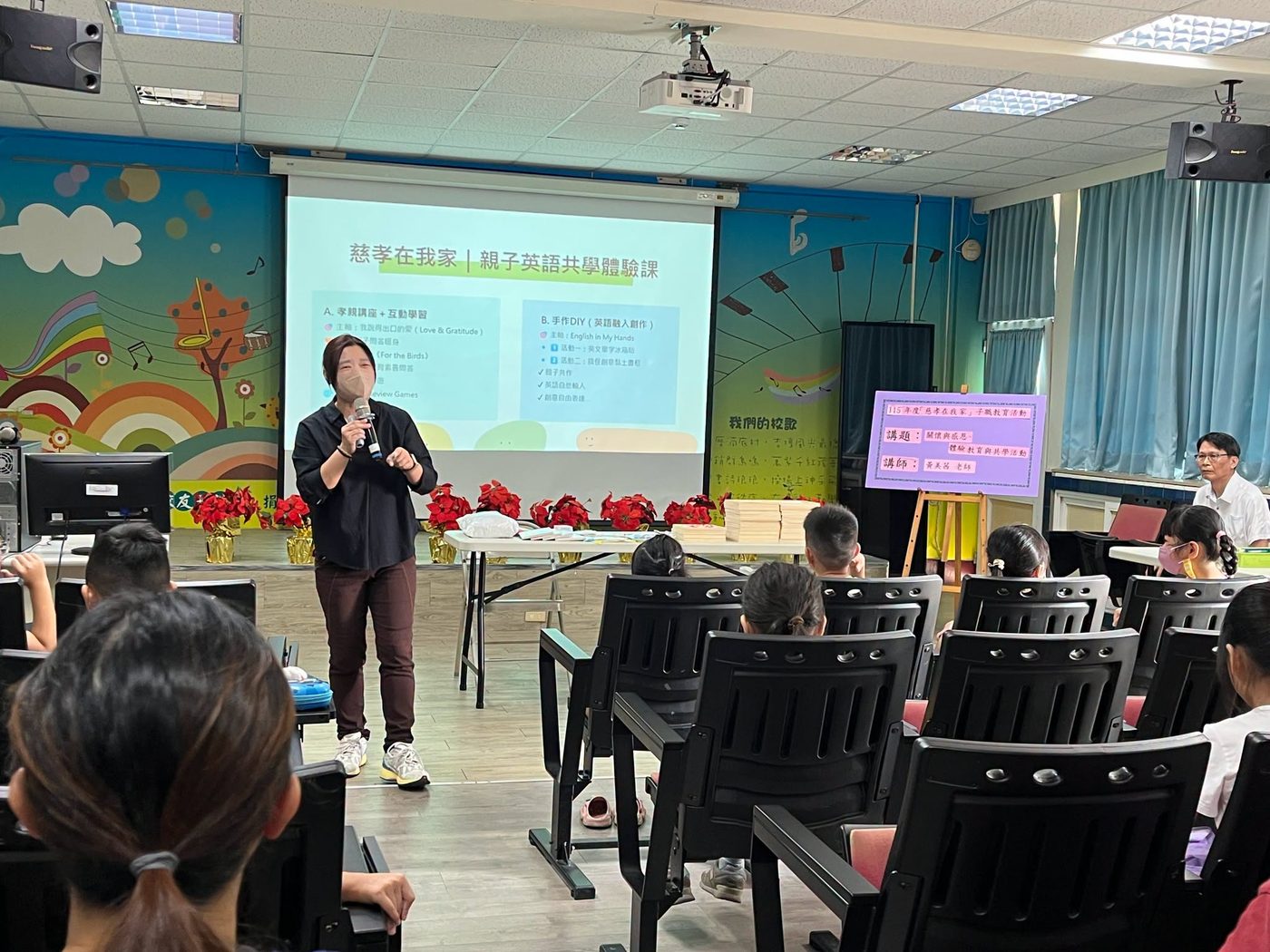 黃美茜老師在橋頭國小視聽教室授課：老師拿著麥克風站在投影幕前，幕上標題寫著「慈孝在我家｜親子英語共學體驗課」，下方分成兩欄，左欄「A. 孝親講座＋互動學習」寫著主軸「我說得出口的愛（Love & Gratitude）」、親子問答暖身、故事《For the Birds》與 Review Games，右欄「B. 手作DIY（英語融入創作）」寫著主軸 English in My Hands、活動一英文單字冰箱貼、活動二搞怪創意黏土畫框，以及親子共作、英語自然輸入、創意自由表達；台下坐著親子，牆面是彩虹與樹木的彩繪 The guest teacher at work in the audiovisual room at Qiaotou — a teacher with a microphone in front of a slide titled 慈孝在我家｜親子英語共學體驗課, split into two panels: on the left the gratitude talk under the heading Love & Gratitude with a warm-up question round, a story and review games; on the right the hands-on session under the heading English in My Hands, listing word magnets and clay picture frames, with parents and children seated below against a wall painted with rainbows and trees