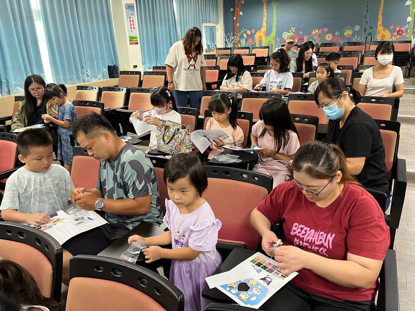 橋頭國小親子一起製作英文單字冰箱貼：視聽教室的座椅上，家長與孩子低頭用剪刀沿著印好的英文單字卡剪下圖案，桌板上放著講義與小夾鏈袋，前排一位爸爸與男孩合看一張彩色字卡，右側一位媽媽正拿著剪刀修邊，後排幾組親子也在各自忙著 Parents and children making English word magnets at Qiaotou — families bent over their seat-top desks cutting printed English word cards apart with scissors, handouts and small zip bags beside them, a father and his son sharing one colored card in the front row, a mother trimming an edge at the right, and more pairs working away in the rows behind