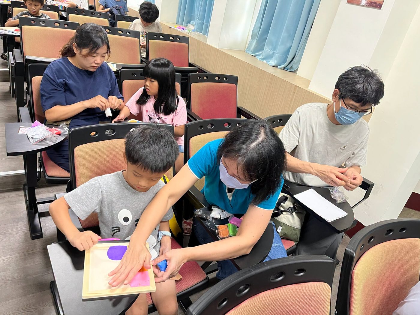 橋頭國小親子共作黏土畫框：一位穿藍綠色上衣、戴口罩的媽媽俯身伸手，協助前座的男孩把一塊藍色黏土按到木頭相框上，框面上已經貼了紫色與粉紅色的黏土色塊；後方另一組母女也在座位上捏黏土，右側一位戴眼鏡、戴口罩的爸爸正低頭揉著手上的黏土 A parent-and-child clay frame in the making at Qiaotou — a mother in a teal top leaning forward to help the boy in front of her press a piece of blue clay onto a wooden frame already covered with patches of purple and pink, another mother and daughter shaping clay in the row behind, and a father in glasses and a mask working a piece between his fingers at the right