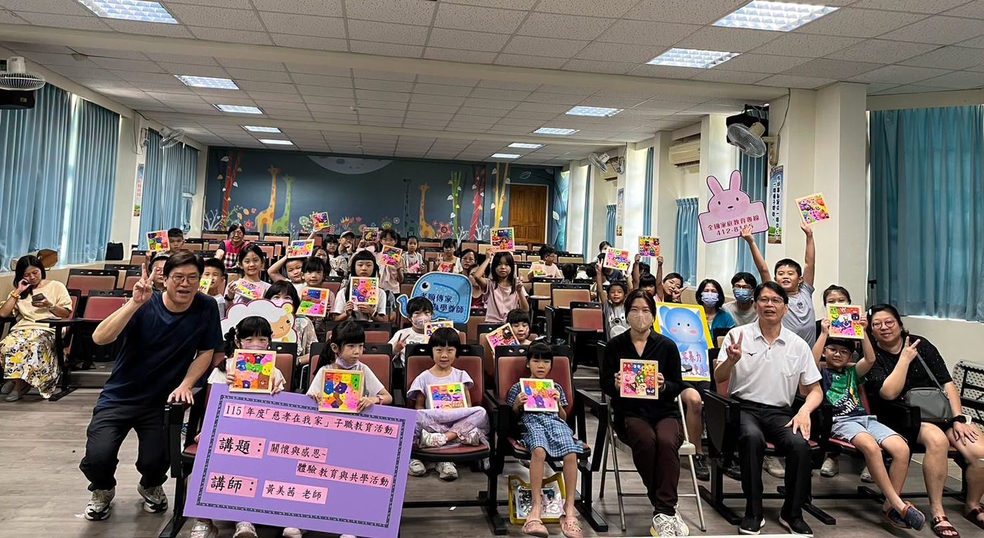 橋頭國小「慈孝在我家」子職教育活動大合照：視聽教室紅色座椅上坐滿孩子與家長，幾乎每個人都把自己剛完成的黏土畫框舉到胸前或頭頂，框裡是五顏六色的黏土小圓點與笑臉；畫面左前方一位穿深藍上衣的男士彎身比出勝利手勢，身旁立著一面紫色海報，寫著「115年度『慈孝在我家』子職教育活動」「講題：關懷與感恩～體驗教育與共學活動」「講師：黃美茜 老師」；右側有人高舉一面粉紅兔子造型的牌子，印著全國家庭教育專線；背景是長頸鹿與花草的彩繪牆 The closing group photograph at Qiaotou — rows of red seats filled with children and parents, nearly every one of them holding up a clay picture frame dotted with colored faces, a man in navy crouching at the front with a victory sign beside a purple board announcing the filial-education day, its topic and its speaker, a pink rabbit-shaped sign carrying the national family-education helpline raised at the right, and a wall painted with giraffes and flowers behind