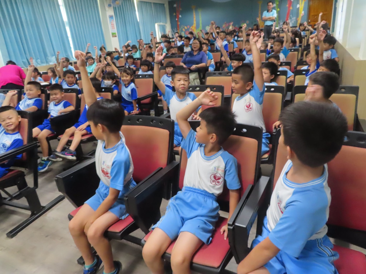 橋頭國小學生在講座中踴躍舉手：視聽教室的紅色座椅上坐滿穿藍白運動服的孩子，前排幾位男孩把手高高舉起，後排幾十隻手也跟著舉，兩位老師站在走道旁，牆上是長頸鹿與彩色樹木的彩繪 Hands up at Qiaotou — rows of red seats filled with children in blue-and-white PE kit, several boys in the front row stretching a hand as high as it will go and dozens more raised behind them, two teachers standing at the side of the aisle, and a wall painted with giraffes and colorful trees