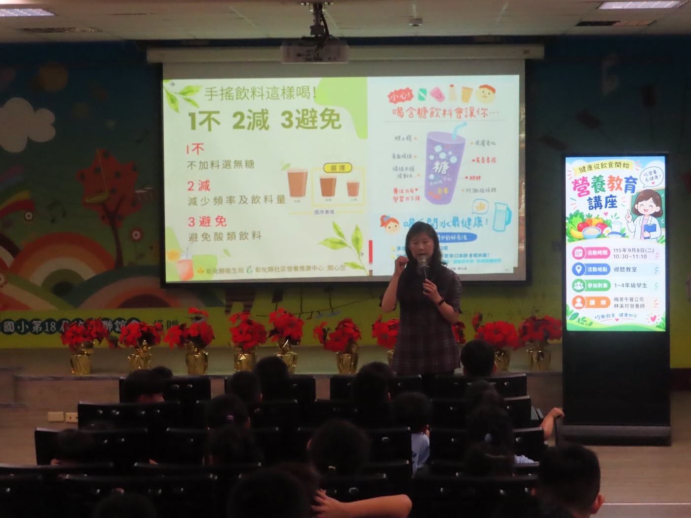 橋頭國小視聽教室的營養教育講座：一位穿格紋上衣的女營養師站在舞台上拿著麥克風講課，身後的大投影幕左半邊寫著「手搖飲料這樣喝！1不 2減 3避免」，並列出「不加料選無糖」「減少頻率及飲料量」「避免酸類飲料」與三杯由大到小的飲料圖；右半邊是一杯紫色手搖飲的插圖，四周標著蛀牙、皮膚老化、長不高、肥胖等字樣，下方寫著「喝白開水最健康！」；舞台右側立著一面電子海報，寫著「健康從飲食開始 營養教育講座」、活動時間 115 年 9 月 8 日（二）10:30~11:10、地點視聽教室、參加對象 1~4 年級學生；舞台前緣排著一整列包著金色紙的聖誕紅盆花，台下坐滿學生 A nutrition talk in the audiovisual room at Qiaotou — a dietitian in a checked top speaking into a microphone on stage, a wide projection screen behind her headed 手搖飲料這樣喝！with the rule 1不 2減 3避免 spelled out beside three cups in descending sizes, and on the right a drawing of a purple bubble-tea cup ringed with the words for tooth decay, aging skin, stunted growth and obesity above the line 喝白開水最健康; a digital poster stands at the right of the stage announcing the talk, its date of September 8, its place and its audience of grades one to four; a row of gold-wrapped poinsettias lines the front of the stage and the seats below are full of children