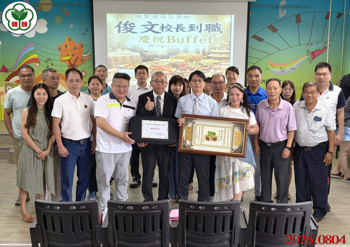 橋頭國小歡迎校長到職餐會大合照：二十多位來賓、家長與教職員在投影幕前排成兩排合影，前排中間兩位男士各捧著一件賀禮&mdash;&mdash;一塊黑色鑲字的匾額與一幅木框裝裱的書法作品，投影幕上寫著「俊文校長到職 慶祝Buffet」，畫面左上角有橋頭國小校徽，右下角有紅色日期 2026.0804 A large group photo at the welcome lunch — more than twenty guests, parents and staff in two rows in front of the projection screen, two men at the front center each holding a congratulatory gift, one a black lettered plaque and one a framed piece of calligraphy, the screen behind reading 俊文校長到職 慶祝Buffet, the school crest watermarked at the top left and the date 2026.0804 in red at the lower right
