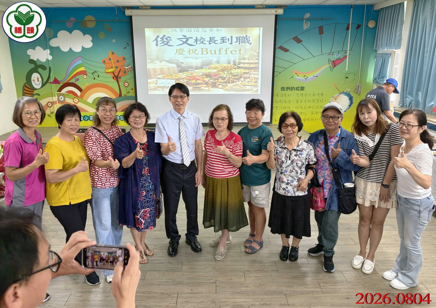 橋頭國小歡迎校長到職餐會合影：十位女士與中間穿淺藍襯衫的校長並排站在投影幕前，大家比出讚的手勢，背景牆面彩繪著音樂主題的彩虹、樂器與白雲，投影幕上寫著「俊文校長到職 慶祝Buffet」，畫面左上角有橋頭國小校徽，右下角有紅色日期 2026.0804 A group at the welcome lunch — ten women standing with the principal in his pale blue shirt in the middle, all giving a thumbs-up, in front of a wall painted with a music-themed rainbow, instruments and white clouds, the projection screen reading 俊文校長到職 慶祝Buffet, the school crest watermarked at the top left and the date 2026.0804 in red at the lower right