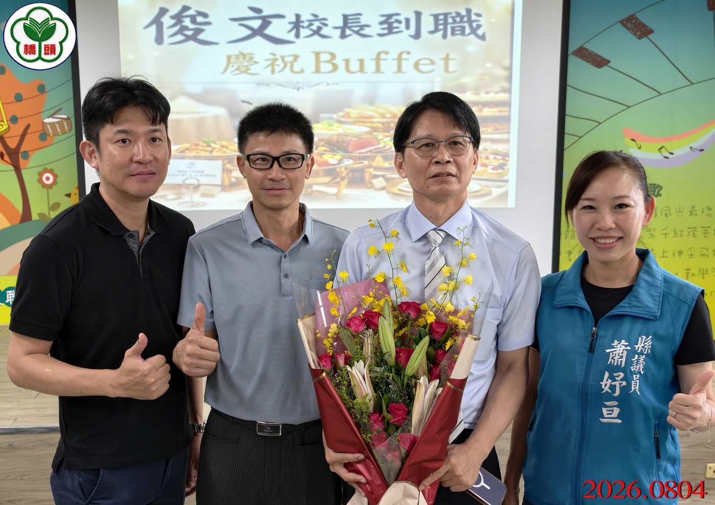 橋頭國小歡迎陳俊文校長到職餐會：室內舞台前，四位大人並排合影，中間的校長穿淺藍襯衫打領帶、雙手捧著一大束紅玫瑰與黃色文心蘭；他左邊兩位男士比出讚的手勢，右邊一位女士穿著繡有「縣議員」字樣的藍綠色背心；背後投影幕上寫著「俊文校長到職 慶祝Buffet」並印著自助餐檯的照片，畫面左上角有橋頭國小校徽，右下角有紅色日期 2026.0804 The welcome lunch for Principal Chen at Qiaotou — four adults side by side in front of an indoor stage, the principal in the middle in a pale blue shirt and tie holding a large bouquet of red roses and yellow orchids, two men giving a thumbs-up at his left and a woman at his right in a teal vest embroidered 縣議員, a projection screen behind reading 俊文校長到職 慶祝Buffet over a photograph of a buffet table, the school crest watermarked at the top left and the date 2026.0804 in red at the lower right