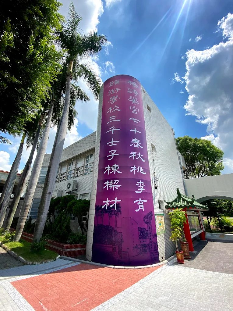 橋頭國小的校慶紀念柱：一根圓柱形的建築量體漆成由粉紫到深紫的漸層，柱面以白色毛筆字直書一副對聯，右聯寫著「橋跨黌宮七十春秋桃李育」，左聯以「頭」字起首，柱身下段以單色印出校園建築與椰子樹的線稿，柱旁是幾棵高大的大王椰子與一座綠瓦紅柱的涼亭，天空湛藍有雲 The anniversary column at Qiaotou — a cylindrical volume of the building painted in a gradient from pale to deep purple, a couplet running down it in white brush calligraphy with the right-hand line reading 橋跨黌宮七十春秋桃李育 and the left-hand line beginning with 頭, a monotone line drawing of the school buildings and palms printed across its lower half, tall royal palms and a green-tiled pavilion with red posts beside it, under a deep blue sky