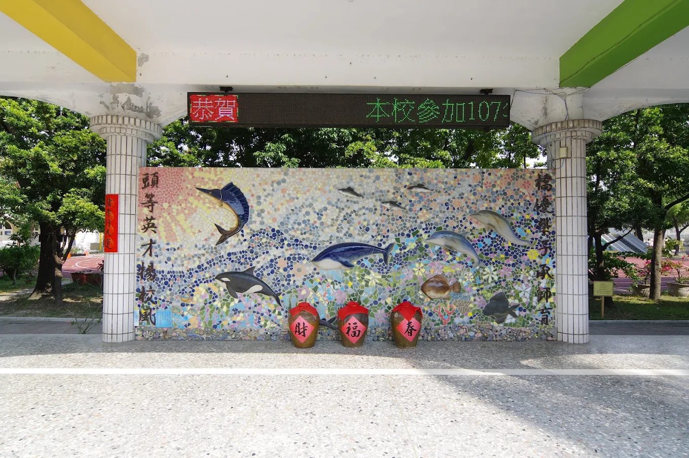 橋頭國小穿堂的馬賽克照壁：兩根白色磁磚方柱之間鑲著一整面碎磁磚拼貼的海洋圖畫，旗魚、海豚、虎鯨與比目魚在藍色海浪中游動，上方拼出旭日光芒；左右柱面各以毛筆字直書一句對聯，牆前擺著三只寫著「財」「福」「春」的陶甕，柱上方掛著一台紅綠雙色的 LED 跑馬燈 The mosaic screen wall in the entrance hall at Qiaotou — a whole seascape in broken tile set between two white-tiled columns, with a sailfish, dolphins, an orca and a flatfish swimming through blue waves and a rising sun radiating above, a line of brush calligraphy running down each column, three ceramic jars lettered 財 福 春 standing in front, and a red-and-green LED ticker mounted overhead