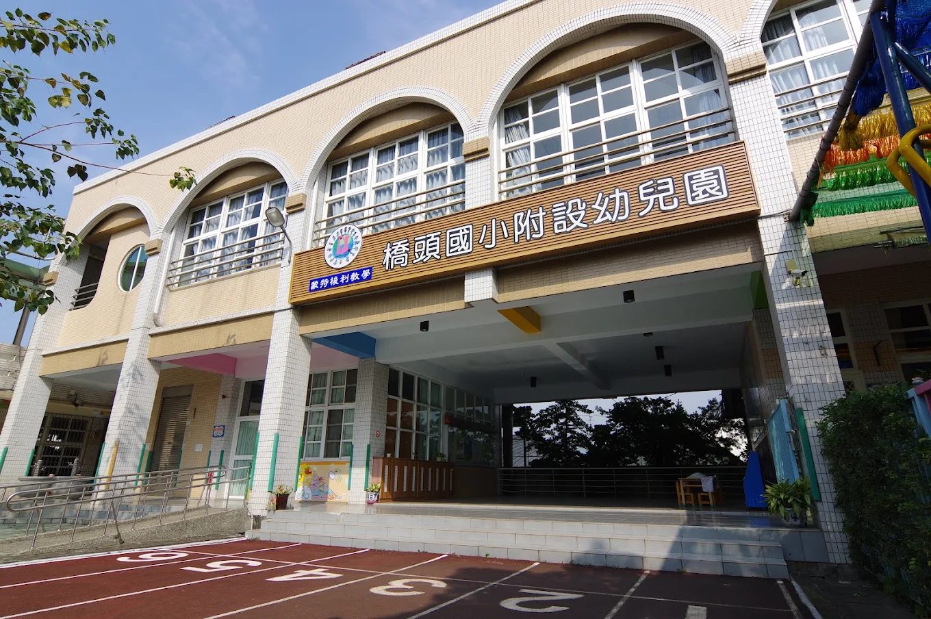 橋頭國小附設幼兒園：一棟兩層樓的米色磁磚建築，二樓有三道白框拱形大窗，窗下掛著一面深色木質招牌，白字寫著「橋頭國小附設幼兒園」，左端另有一小塊藍底招牌；一樓是挑高的開放門廊，地面延伸出紅色跑道與白色數字標線，右側掛著彩色的裝飾旗幟 The kindergarten at Qiaotou — a two-storey cream-tiled building with three tall white-framed arched windows on the upper floor, a dark timber board beneath them lettered in white 橋頭國小附設幼兒園 with a small blue sign at its left end, an open double-height porch at ground level, red track surfacing and white lane numbers running up to it, and colored bunting hanging at the right