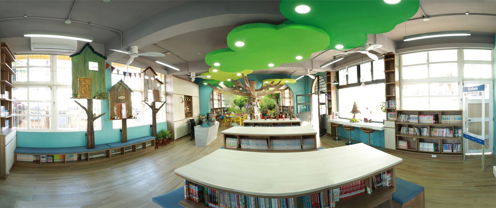 橋頭國小的閱讀室內部環景：天花板懸掛著綠色雲朵造型的燈板，中央立著一棵造型樹，牆邊做出木屋與樹幹造型的裝飾與座椅，湖水綠的牆面搭配木紋地板，中間是弧形的木質書架與閱讀桌，四周是滿櫃的童書與大片採光窗 The reading room at Qiaotou seen in a wide panorama — green cloud-shaped light panels hanging from the ceiling, a sculpted tree in the middle of the room, timber cabin and tree-trunk shapes built along the walls with bench seating, pale teal walls above a wood-grain floor, curved wooden bookcases and reading tables in the center, and shelves of children's books beneath tall windows