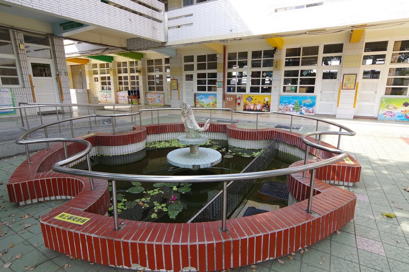 橋頭國小中庭的生態池：一座紅磚砌成的環形水池，池緣圍著不鏽鋼扶手，池中央的石台上立著一尊躍起的魚形雕塑，水面浮著睡蓮與一朵粉紅色的花，池後方是掛滿彩色海報的教室走廊 The courtyard pond at Qiaotou — a ring-shaped pool built in red brick with a stainless-steel handrail around it, a leaping-fish sculpture standing on a stone plinth at its center, water lilies and a single pink bloom floating on the surface, and the classroom corridor hung with brightly colored posters behind
