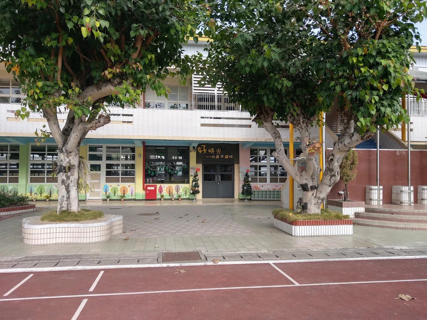 橋頭國小的社區共讀站入口：兩棵樹幹粗大的老榕分立左右，樹穴砌成八角形的白色磁磚花台，正中央是一間玻璃門的教室，門楣上掛著木質招牌，橘色大字寫著「好時光」，下方小字寫著「社區共讀站」，牆面下段漆成粉綠與粉紅，門旁擺著一座紅色郵筒與一整排彩繪的樹形立牌，前方是紅色的 PU 地坪與白色球場線 The community reading station at Qiaotou — two thick-trunked old banyans on either side standing in octagonal white-tiled planters, a glass-doored room in the middle with a timber sign above the lintel reading 好時光 in large orange characters and 社區共讀站 beneath, pale green and pink panels along the lower wall, a red postbox and a row of painted tree-shaped boards beside the door, and red paving with white court markings in front