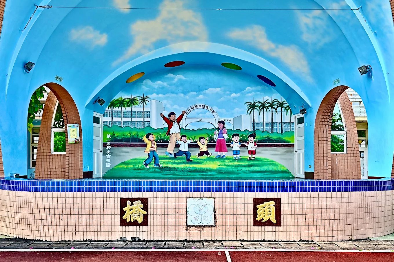 橋頭國小司令台正面：一座漆成天藍色的半圓形拱頂舞台，拱頂內畫著白雲，背板上畫著七個孩子在綠色草地上奔跑跳躍、一位老師張開雙手站在中間，畫面背景是校舍與椰子樹；台座貼著米色與藍色磁磚，正面由左至右嵌著金色的「橋」字、校徽浮雕與「頭」字 The stage at Qiaotou seen head-on — a semicircular sky-blue canopy painted inside with white clouds, a backdrop mural of seven children running and leaping on green grass with a teacher standing among them arms wide, school buildings and coconut palms painted behind them, and a tiled apron below carrying a gold 橋, a relief of the school crest, and a gold 頭