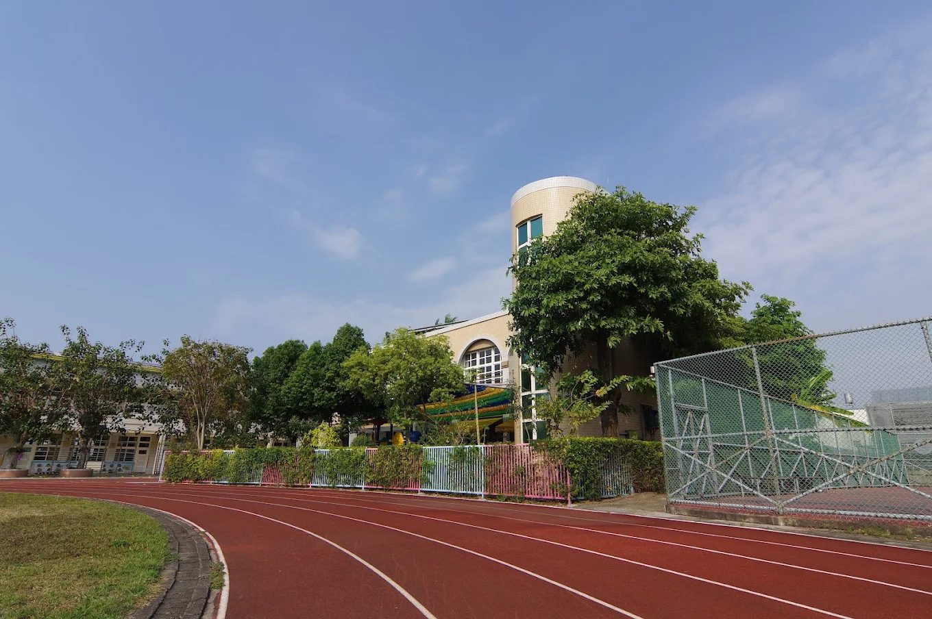 橋頭國小的跑道與圓塔：紅色 PU 跑道由前景往左後方彎去，右側是綠色鐵絲網球場，跑道內側種著一排修剪成球形的樹與粉藍相間的欄杆，正後方是一棟米色校舍，屋角是一座圓柱形的塔樓，塔身開著窄長的窗，旁邊一棵大樹幾乎把塔遮住一半，天空是藍色的 The track and tower at Qiaotou — red synthetic lanes curving away to the left, a green wire-fenced court on the right, a row of ball-clipped trees and a pink-and-blue railing along the inside of the track, and a cream school building behind with a cylindrical tower at its corner, narrow windows up its side and a large tree half covering it, under a blue sky