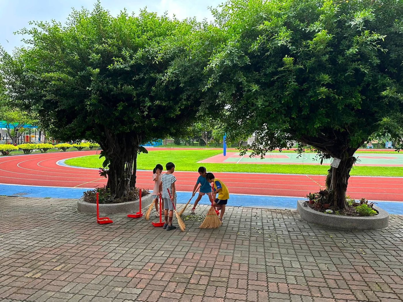 水尾國小返校日,學生在跑道旁的大樹下拿掃把打掃