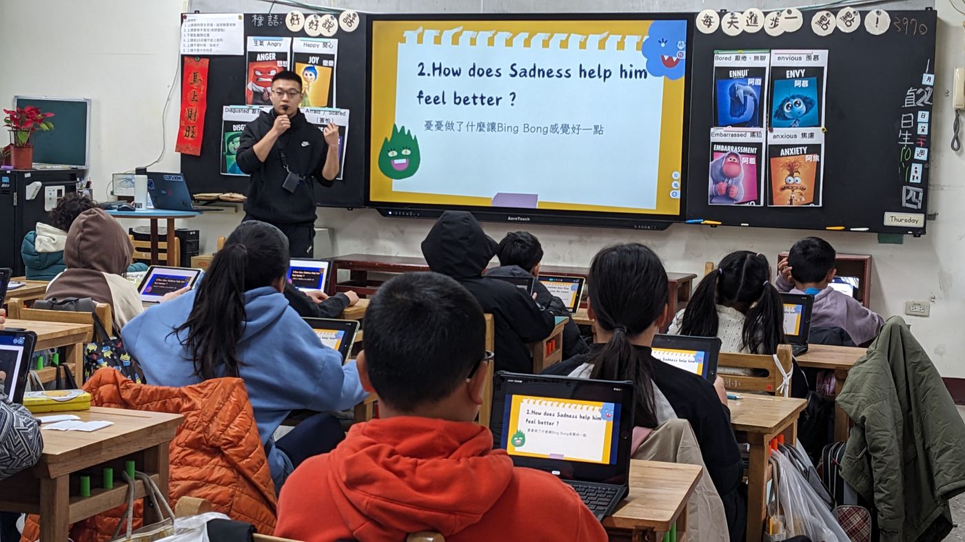 Grade 5 bilingual class — students at desks with tablets, teacher at board with Sadness slide