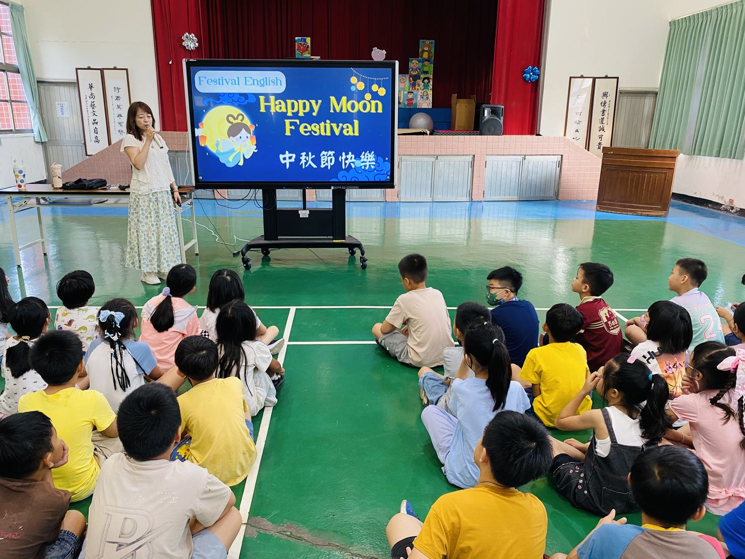 老師站在「Happy Moon Festival 中秋節快樂」投影畫面旁，學生坐在地上舉手 A teacher beside a "Happy Moon Festival" slide as students sitting on the floor raise their hands