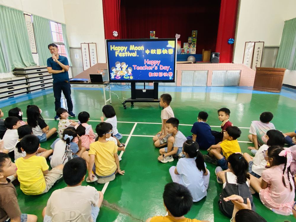 螢幕上寫著 Happy Moon Festival 和 Happy Teacher's Day，全校學生坐在禮堂 The screen showing Happy Moon Festival and Happy Teacher's Day greetings to students in the hall