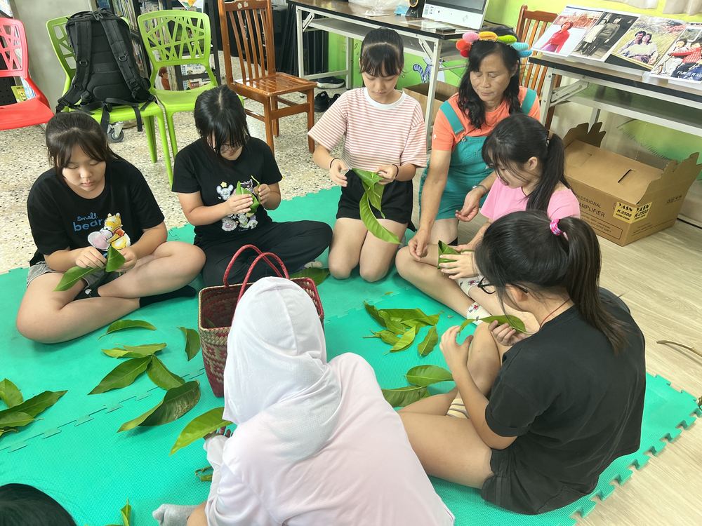 學生坐在地墊上用植物葉子編織童玩 Students sitting on mats, weaving toys from plant leaves