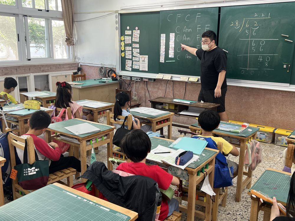 老師在黑板前教烏克麗麗和弦，學生坐著練習 A teacher showing ukulele chords at the blackboard while students practice at their desks
