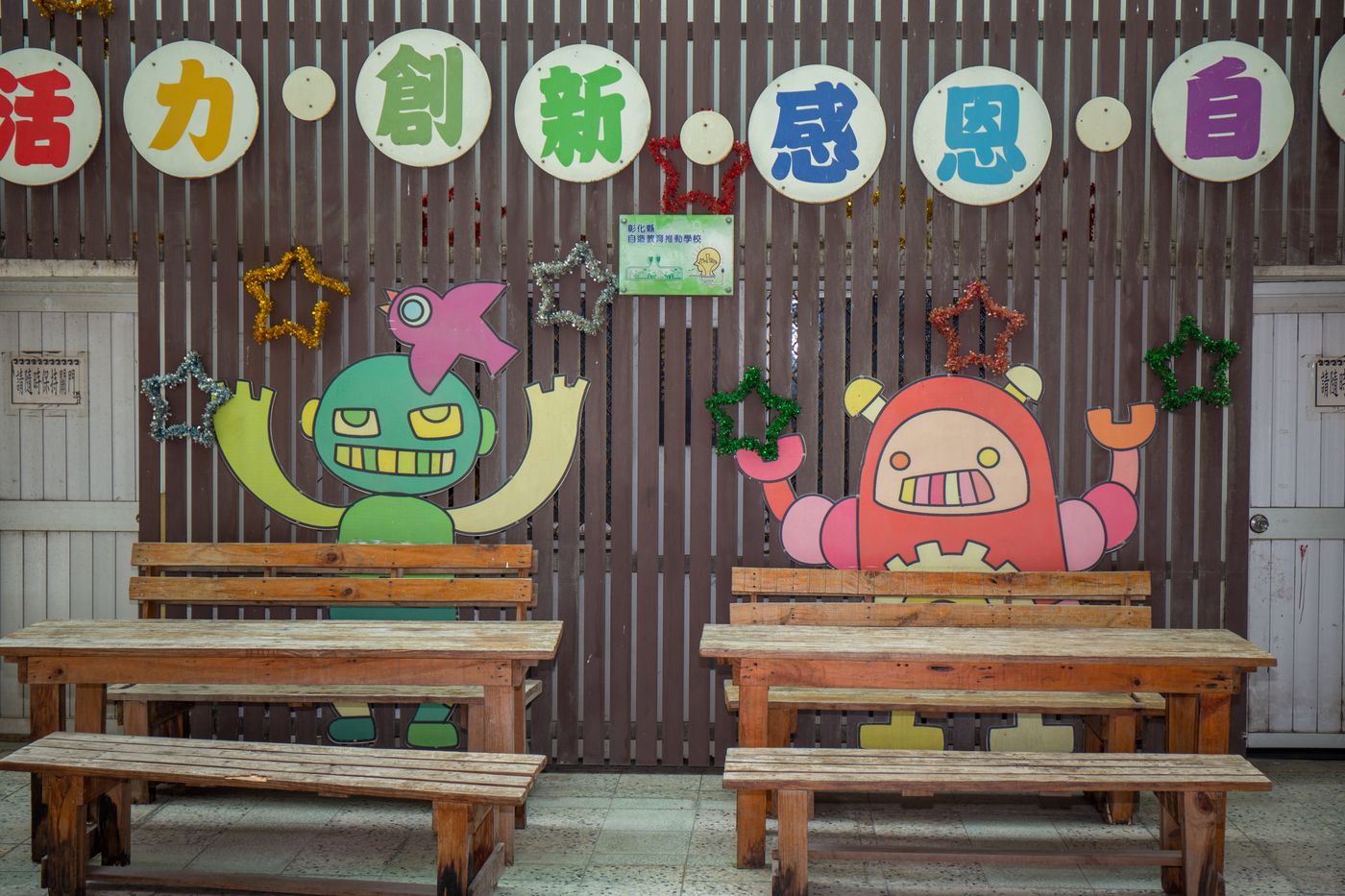 興華國小校園牆面上掛著「活力、創新、感恩、自信」彩色圓牌，前方有兩張木頭長椅與可愛的機器人圖案 A SingHua campus wall with colorful round signs reading 活力, 創新, 感恩, 自信, above two wooden benches and cheerful robot cut-outs