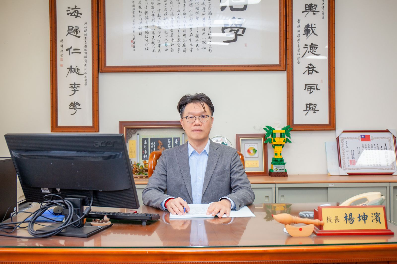 楊坤濱校長坐在校長室辦公桌前，身後掛著書法作品，桌上立著「校長 楊坤濱」名牌 Principal Yang Kun-bin at his desk in the principal's office, with calligraphy scrolls behind him and his nameplate on the desk