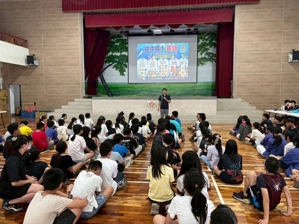 田中國小活動中心舞台大螢幕顯示「田中國小英語 TJES English Day」活動看板，全校學生席地而坐