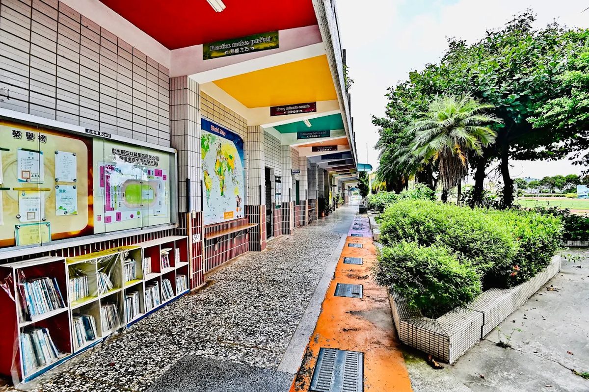 萬合國小的教室走廊：左側是開架書櫃與玻璃公佈欄，牆上掛著一張世界地圖，走廊天花板一段一段漆成紅、黃、綠、藍，橫樑下懸著英文標語木牌，右側是樹籬與棕櫚樹 A classroom corridor at Wanhe — open bookshelves and a glazed noticeboard on the left, a world map on the wall, the ceiling painted in successive bands of red, yellow, green and blue, English mottoes hanging from the beams, and hedges and palms along the right