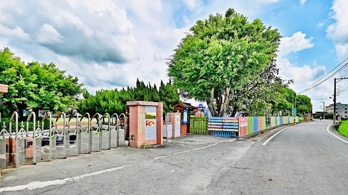 萬合國小校門外的鄉道：柏油路沿著學校延伸，左側是不鏽鋼校門與白色欄杆，牆邊立著一排彩色木條圍籬，門內可見遊具與跑道，兩旁是老樹與農村住宅，天空藍白相間 The country road outside the Wanhe gate — asphalt running along the school boundary, the stainless-steel gate and white railings on the left, a run of brightly painted picket fencing along the wall, play equipment and the track visible inside, old trees and village houses on either side under a part-cloudy sky